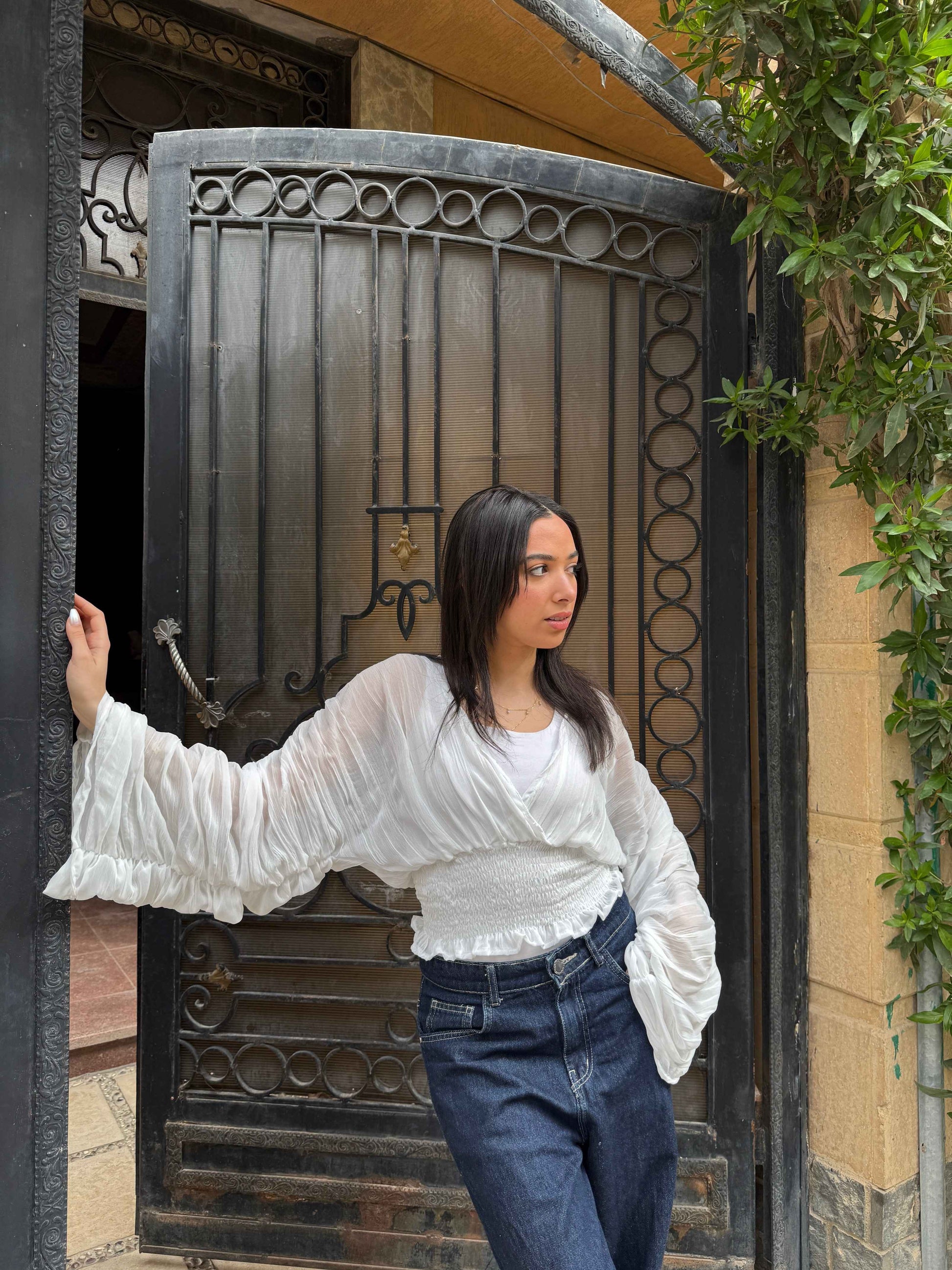 Ocean Breeze Blouse – elegant white pleated blouse with flowing dramatic sleeves and a smocked waist, styled casually with dark denim in an outdoor urban setting.