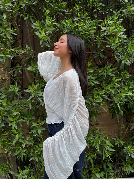 Ocean Breeze Blouse – woman wearing a flowing white textured blouse with dramatic pleated sleeves, posing naturally outdoors against lush green foliage.